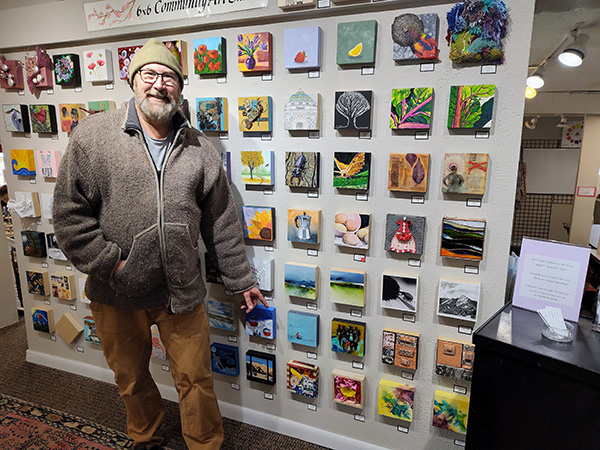 Wall full of 6"x6" paintings with Eric Howell pointing out his and Angela Alexander's paintings